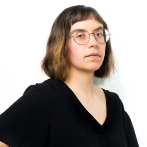 This image is a headshot of 2023 creative glass fellow Paige Lizbeth Morris, who is standing with her hand on her hip, wearing round glasses, and has short brown hair.