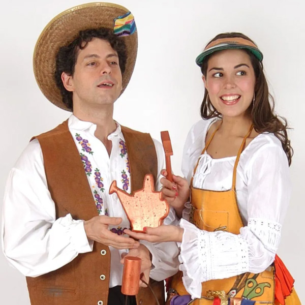 2 actors in the NJ Theatre Alliance's Stages Festival are smiling and wearing costumes while holding small props together. The left actor is a man wearing a white long sleeve shirt, a vest, and a straw hat. To the right is a woman with long brown hair, a white dress, and an orange apron.