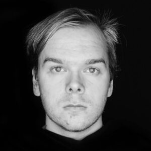 A grey-scale headshot of 2023 creative glass fellow Matt Jacob, who is looking directly at the camera and wearing a dark sweatshirt, is the image's subject.