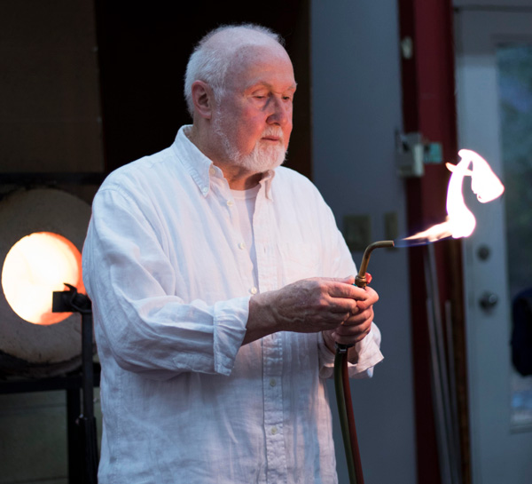 This image is a medium close-up of Paul Stankard, wearing a light blue button-up, and holding a torch.