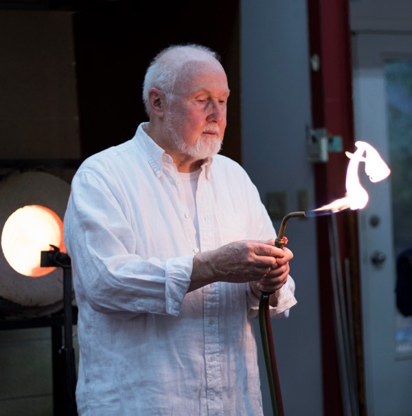 This image is a medium close-up of Paul Stankard, wearing a light blue button-up, and holding a torch.