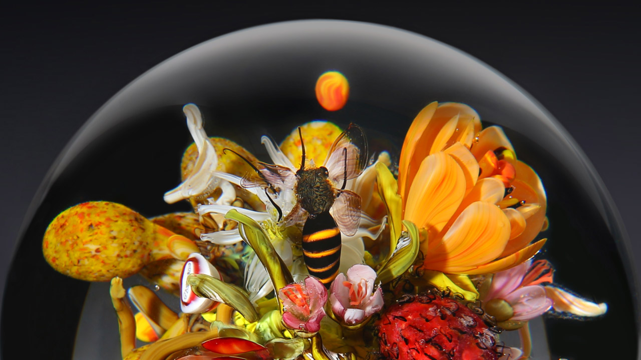 This banner shows an image of the top of a paperweight with a glass bee miniature sitting among various bright-colored glass flowers.