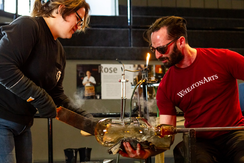 Image of two WheatonArts glass artists. One is wearing a red t-shirt that reads "WheatonArts" and is sitting on a workbench that has a metal rod resting on the railing. Attached at the end of the rod is a large amount of amber colored, bottle-like shaped hot glass. The artist is holding a rag under the glass, while the other artist uses a flat rectangular tool to shape it. Smoke is rising from where the rag meets the glass.