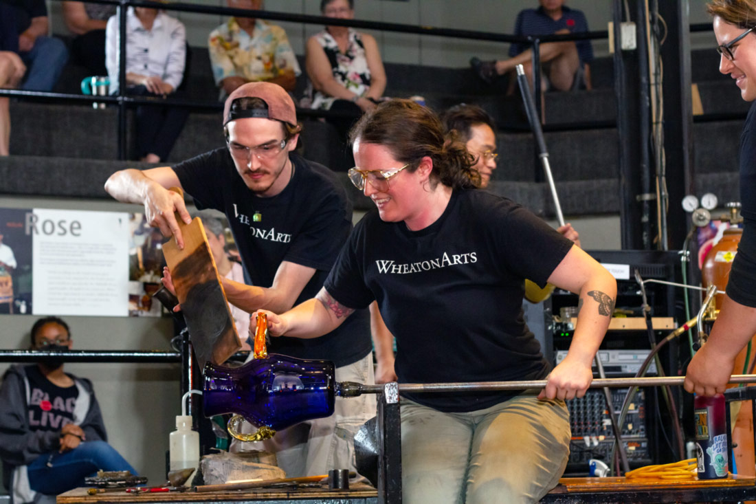This photo is of Glass Studio Coordinator Katey Murphy & assistant Jeremy Hayes shaping a blue glass vase in the Glass Studio. Katey is sitting on a workbench holding the rod with her left hand and an orange tool on her right that she is using on the piece. Jeremy is using a wood plank to shield Katey's arm.