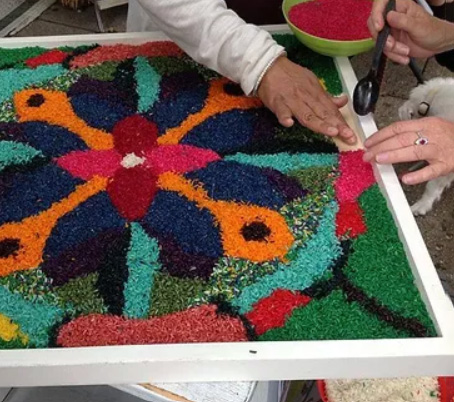 This image is a close-up of the ECO Fair Indian Earth Mandala demonstration with artist Rita Pundyu, presented by a light blue circle. Red & pink border accents are formed with beads by two hands belonging to different people. There is an orange, light blue, dark blue, and pink pattern in the center.
