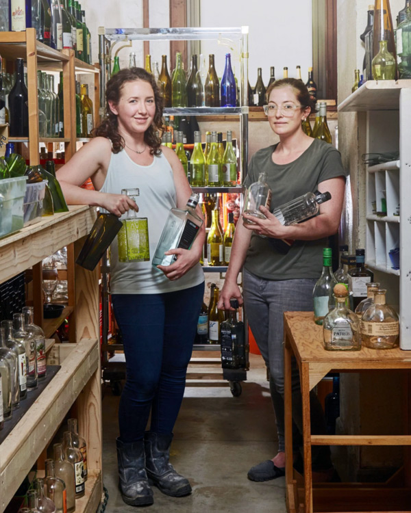 Two women from ECO Fair hot glass demonstrators are a part of Remark Glass. They are holding several glass bottles and are surrounded by racks of glass bottles.