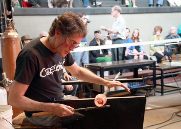 Image of Gordon Smith, during a glass blowing demonstration. Smith is sitting on a glass blowing bench, rolling a metal rod, with orange hot glass at the end, with one hand, while the other hand uses a tool to sculpt the hot glass. A crowd of people sit and watch him from the background.