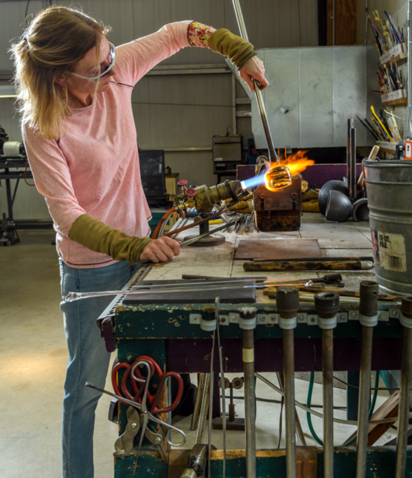 Image of Melissa Ayotte, who is wearing protective glasses, a long sleeved, light pink shirt, light green protective forearm sleeves, and light blue jeans. With one arm raised, Ayotte holds a metal rod and is directing the hot glass, at the end of the rod, towards a lit torch. The torch's light blue flame turns orange as it hits and extends past the glass.
