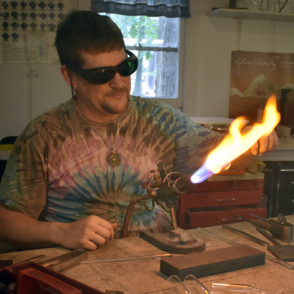 Image of Bill Futer, who is wearing a tie-dye shirt, black glasses, and a spiral pendant. Bill sits in front of a torch and is directing glass towards the flame. The flame starts out blue, but turns orange as it hits the glass.