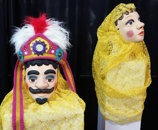 Image of two masks resting on two separate white podiums against a black background. One mask depicts a man with a multicolored headpiece that drapes over the podium. The other mask depicts a woman whose yellow headpiece is draped over the podium.