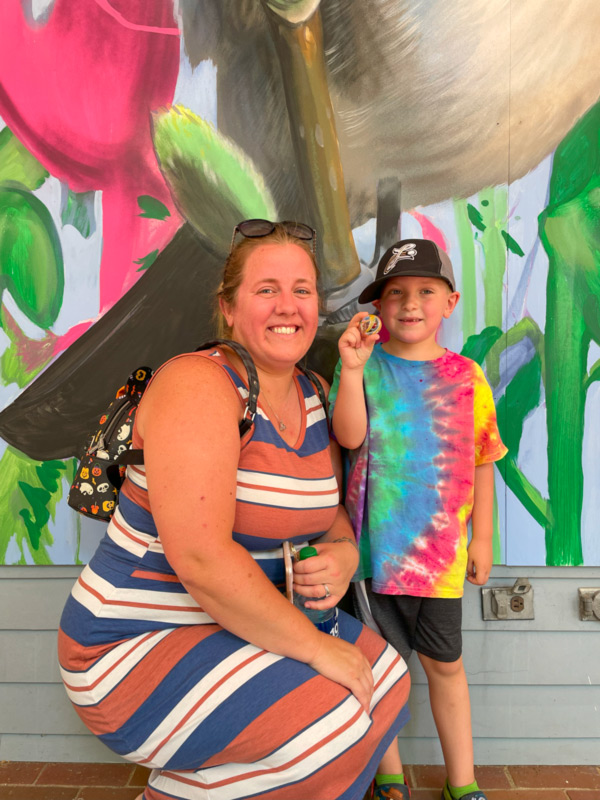Photo of the child who won the 2022 marble hunt. The child is wearing a tie-dye shirt and is holding a marble up. Next to him is an adult crouching down to the child's height. Both individuals are standing in front of a colorful mural and are smiling at the camera.