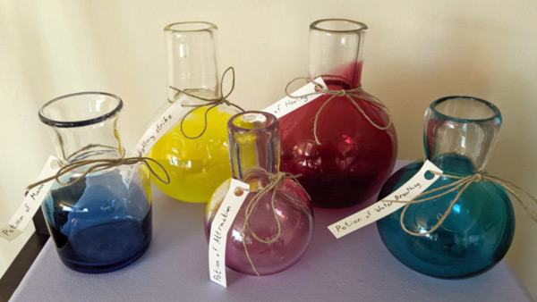 Image of five different colored glass potion bottles of various heights and shapes against a white background. Each bottle has a white label, with a handwritten title detailing the potion's powers.