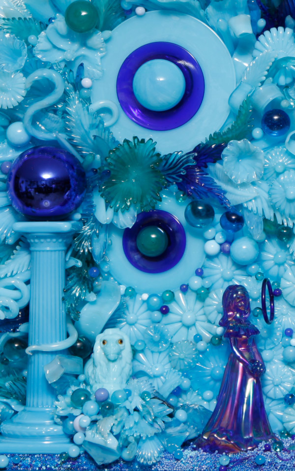 Image from the Amber Cowan exhibit. The piece displays a blue iridescent glass woman, in a long gown with her hands clasped in front of her. She is facing in the same direction as a dark blue ring that is floating in front of her face. To the left of her is a light blue sheep resting in a bushel of glass spheres and flowers that are various shades of blue. To the left of the sheep, is a large light blue podium with a dark blue sphere resting on the top. All of this sits in front of a glass background of spheres, flowers, and leaves that are various shades of blue.