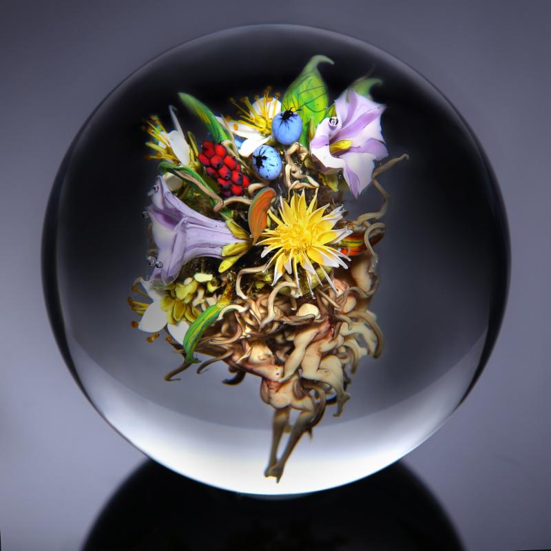 Image of a paperweight by Paul Joseph Stankard. The paperweight is a clear globe on a dark gray background. Inside the orb is a cluster of a variety of red, light purple, white, yellow, and blue flowers with green leaves. Nude bodies are at the bottom of the cluster and curly pieces of the same color extend towards the flowers.