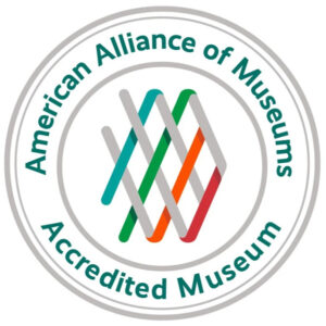 Image of a circular logo that features three circles inside of one another. The first circle has a thick light gray line, but the light gray circle inside of it is thinner. There is dark teal text on the top of the circle, that follows the curve, that reads "American Alliance of Museums". There is text of similar style at the bottom, that reads "Accredited Museum". This is followed by another thick light gray circle. In the center of the circle are four diagonal thick light gray lines that are woven with four thick diagonal teal, green, orange, and maroon lines that are angled oppositely of the gray lines.