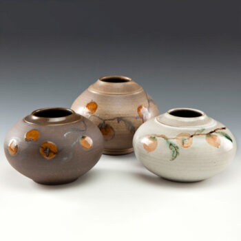 Image of three short and wide clay vases in front of a white to dark gray gradient background. The vase on the left is brown with orange fruit on a dark leafy vine across the middle. The middle vase is the same, but is a light brown. The vase on the right is also the same, but is white.