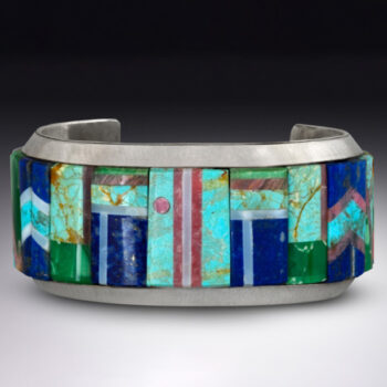 Image of a thick silver ring in front of a white-to-black gradient background. In the middle of the band are rectangular sections of different light blue, dark blue, green, and pink stone designs.
