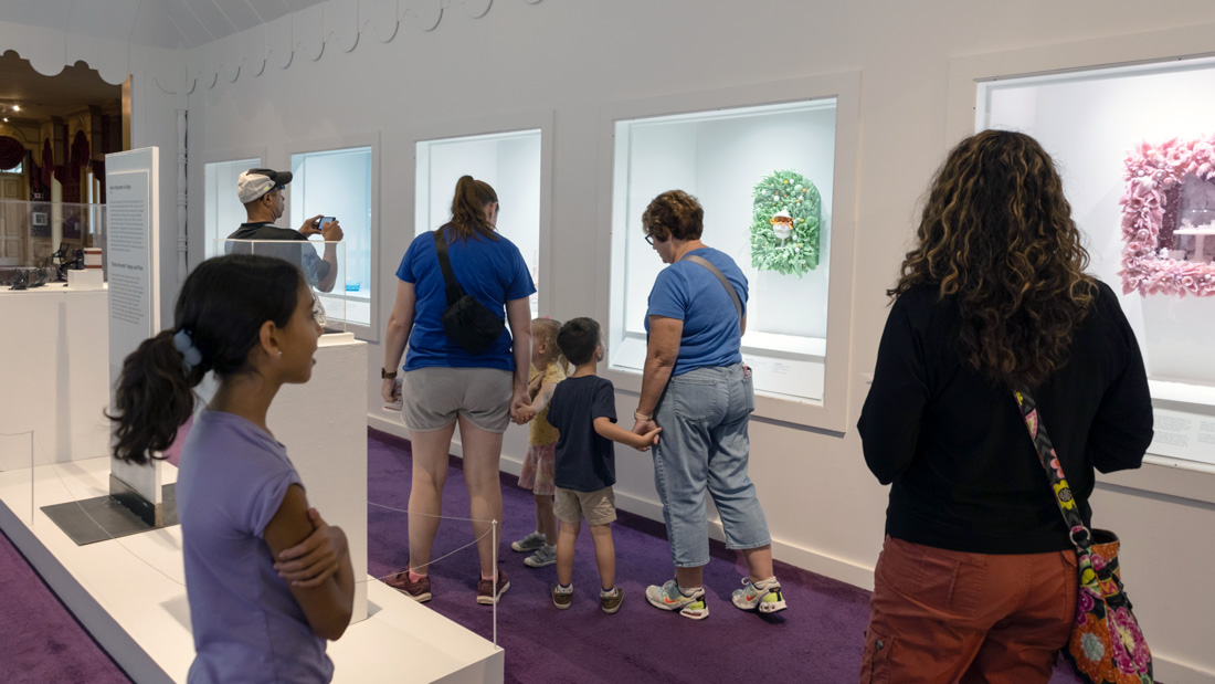 Four adults and three children in a rectangular room within the Museum of American Glass. The visitors are all gazing at the pieces from the Amber Cowan Alchemy of Adornment exhibit that are displayed in window cases within the white walls. There are two white podiums with clear cases resting on a rectangular white stand in the center of the room on top of a purple carpet. There are more pieces from the exhibit resting on the podiums. A tall white sign displaying information regarding the exhibit is in between the two podiums. There is a golden doorway that leads to the next room in the far left corner.
