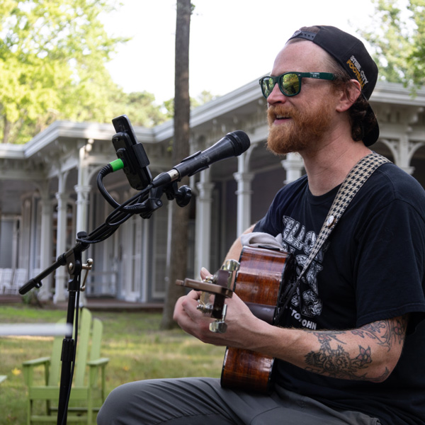 Image of Chris Lax performing outside in front of the Museum of American Glass. Chris is playing a guitar with a black and white houndstooth strap, while sitting in front of and singing into a microphone. Chris has a black hat, black sunglasses, a reddish brown beard, and is wearing a black t-shirt.