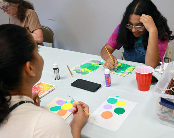 Image of three people participating in the "Community Mural Squares: Art and Nature" Free Family Art Activity held during Wheaton Wednesday. Each person has a pale wooden square in front of them, that they are painting. One of the two pictured squares has a six petal flower in the right corner. When the artist is finished, the flower will have two teal petals, two purple petals, and two dark orange petals. One of the teal petals is still in progress. The flower also has a bright yellow circular center. Next to the flower is a palette filled with the colors being used on the square. On the other side of the square is a Wheaton Wednesday brochure. In the center of the white table, are two small bottles of black and purple paint, a yellow-tipped paint brush, a red plastic cup, and a clear bin filled with supplies. On the other side of the table is another wooden square that has a green, blue, purple, and yellow nature design being painted on the surface. Another palette sits close by with the colors being used on the square inside of it.