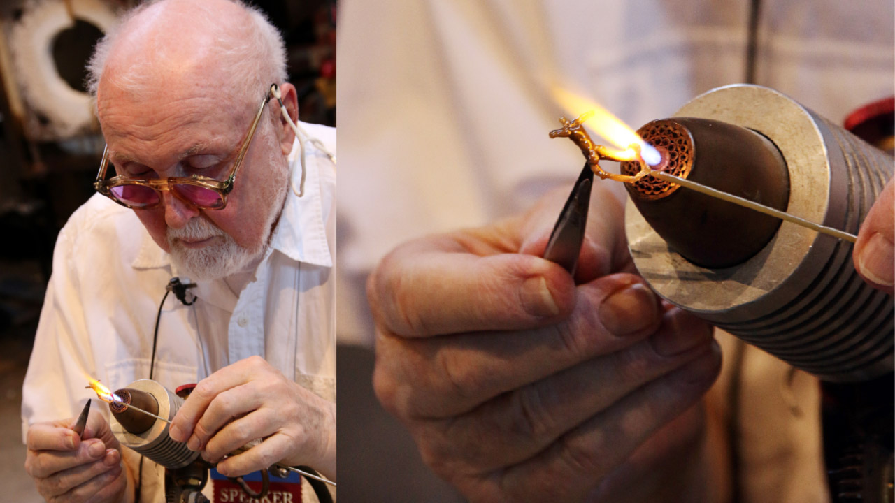 This image is composed of two side-by-side images. The left image shows Paul Stankard, wearing glasses and a white button down shirt. He is working over a torch and is guiding a small flamework piece into the orange flame using two tools. The right larger image is a close up of this process. The image shows Paul holding a small flameworked human body into the flame with the two tools, one in each hand.