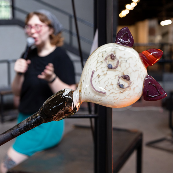 Image of one of the glass masks chosen from the "Design a Glass Mask" contest held during Wheaton Wednesday. The mask is attached to the end of a glass blowing pole. The face of the mask is white with a purple smile and purple circles for eyes. At the top of the mask are two dark purple triangular ears with a short maroon and orange horn in the middle. The image was taken in the Glass Studio, where WheatonArts Glass Artist Kendall Frank is speaking into a microphone in the background.