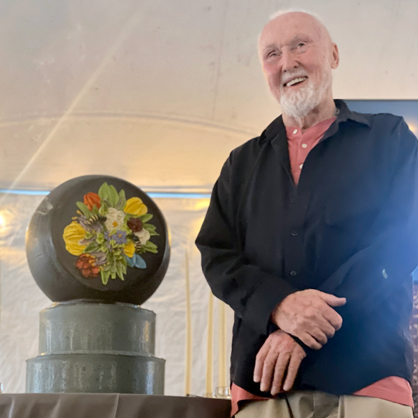 Image of Paul Stankard standing in front of a paperweight cake. The cake paperweight is black with a multicolor floral cluster in the middle. The paperweight sits upon two tiers, which are on top of a white tablecloth. Paul is smiling at the camera and wearing a black button down shirt over top of a light red shirt.