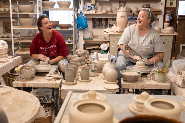 Image of WheatonArts potters Amy Pesseller and Phyllis Seidner-Vesely. Amy and Phyllis are wearing WheatonArts t-shirts and are sitting around two pottery wheels, smiling at each other while shaping clay. They are surrounded by a variety of clay projects.