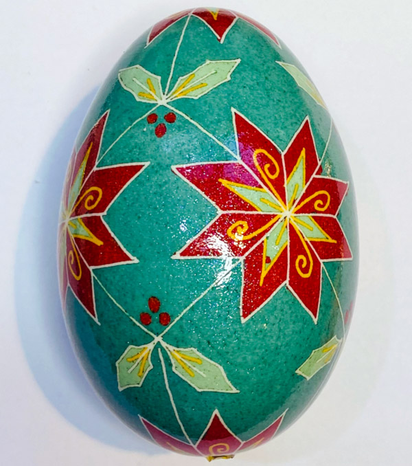 Image of a painted goose egg on a white background. The base of the egg is teal with white lines forming a large diamond pattern. Within the diamonds are red, yellow, and light blue triangular two-layer flowers and leaf designs outlined with white. The diamonds also feature three small red dots in the corners that do not have flower petals or leaves. The other corners of the diamond have either two petals or a leaf. The center of the flower is where the diamonds connect.