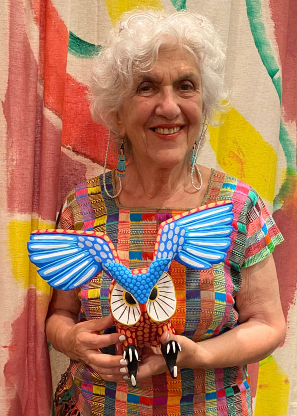 Image of Julia Zagar in front of a colorful background. Julia has short, wavy white hair, a colorful short sleeve shirt, and is holding a brown owl sculpture with white around the eyes, black feet, and blue and white extended wings that start at the owl's forehead.