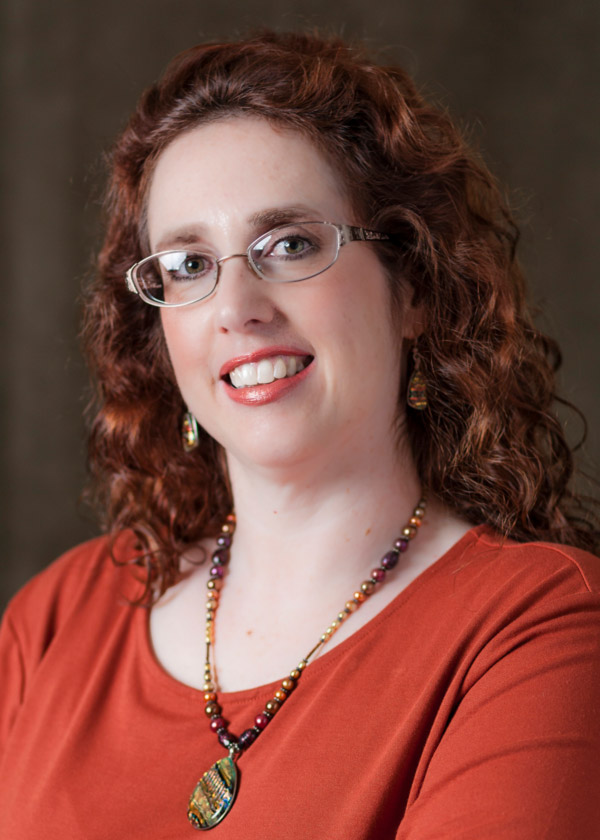 Image of Noele Alampi in front of a black background. Noele has shoulder-length curly brown hair and is wearing rectangular glasses, a brown oval stone necklace that is dangling from a beaded cord, a red long sleeve shirt, and square gold dangling earrings.