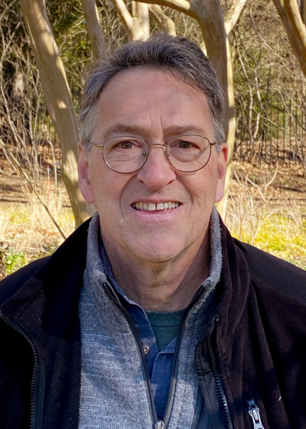 Image of Tony Fisher in front of a wooded background. Tony has short gray hair and is wearing round glasses, a dark unzipped jacket, a light gray sweater with a zipper, a blue button-up shirt, and a dark gray shirt underneath.