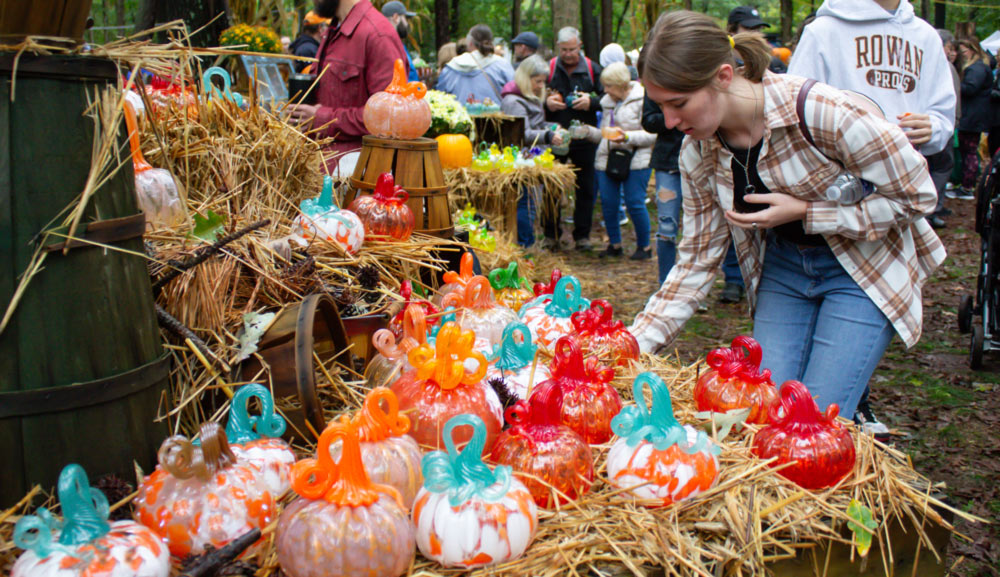 Image of a crowd gathered inside of the WheatonArts Glass Pumpkin Fundraiser. A variety of orange, white, and red glass pumpkins with light blue, red, orange, and green curly stems sit on a wooden surface covered in straw. Wooden baskets and barrels are also featured throughout the set up. Customers are holding and browsing the glass pumpkins. Yellow and blue glass pumpkins can also be seen in the background.