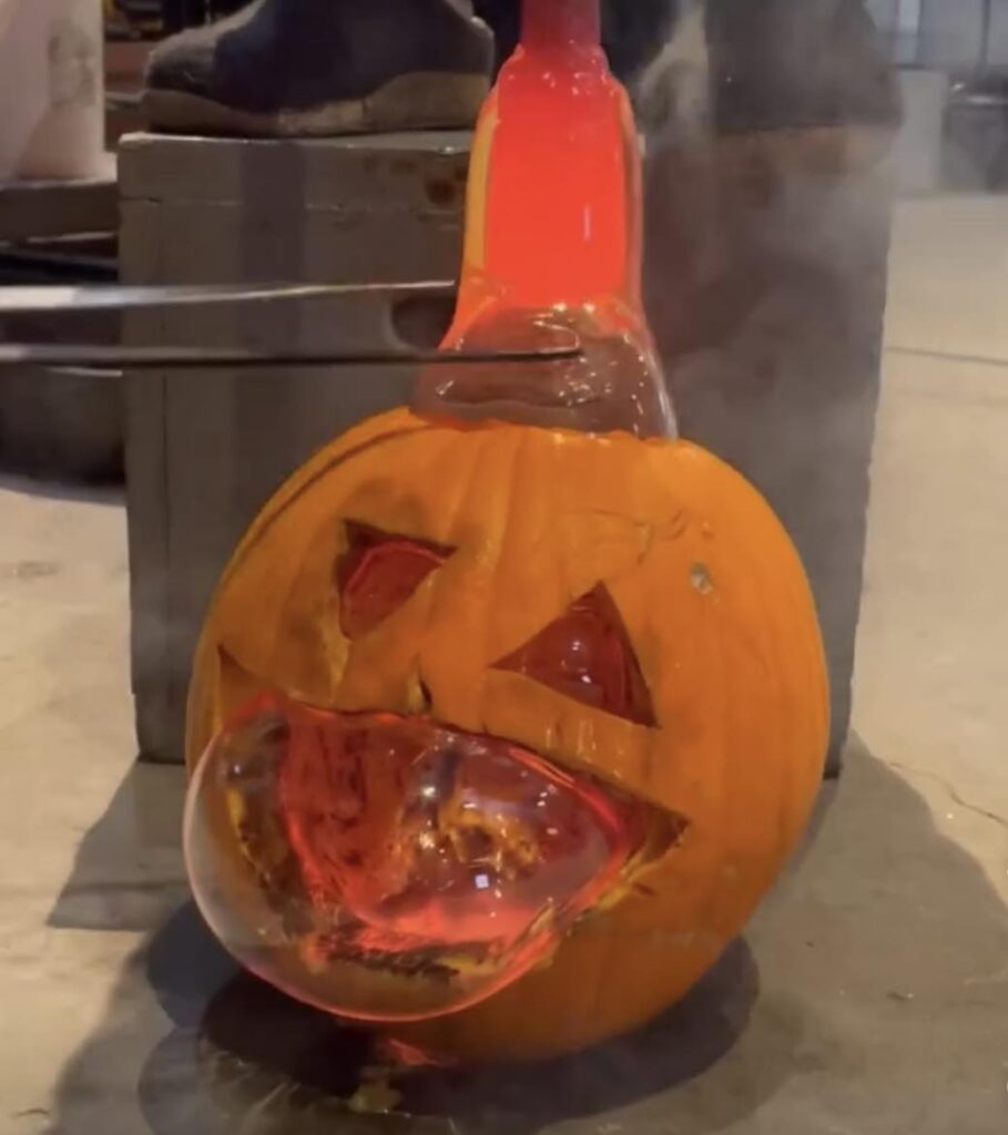 Image of a large orange pumpkin with triangular eyes and a large mouth carved into it. Hot glass is being blown into the top of the pumpkin so that a large glass bubble is coming out of the mouth. A pair on tongs is assisting the glass at the top of the pumpkin.