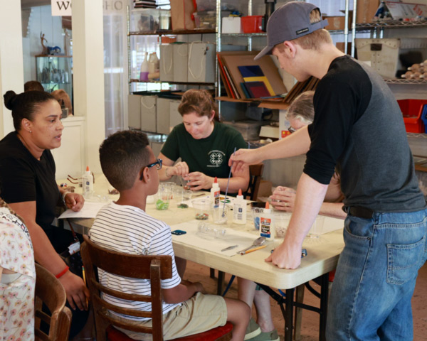 Image of a group of people of all ages sitting around a white table filled with crafting supplies in the WheatonArts Education Studio. One person is standing in front of the table and is dipping a paint brush into a clear plastic cup while the others observe.