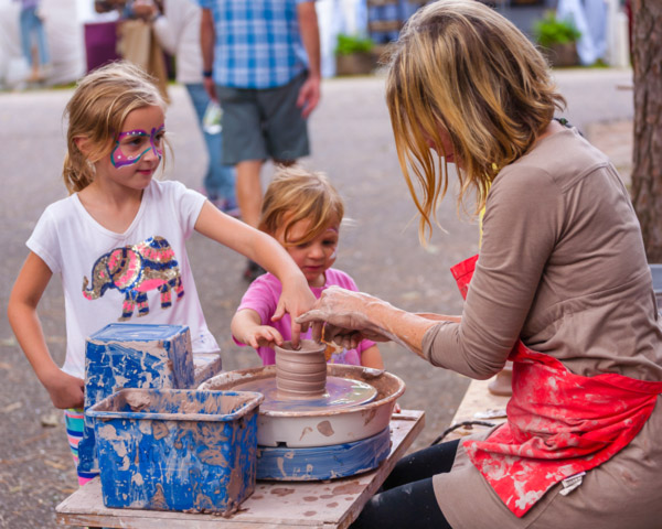 Image of two children assisting a potter outside at the Festival of Fine Craft. Both children are pinching the top sides of the gray clay cylinder on top of a pottery wheel. Each child has face paint and the potter is wearing a red apron. Next to the pottery wheel is two blue bins that are covered in clay.