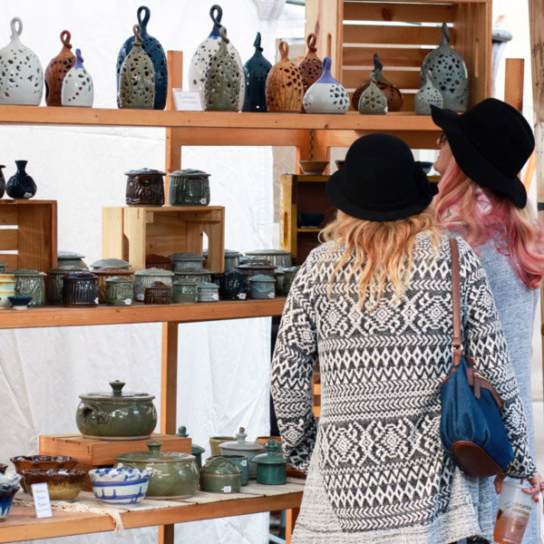Image of two customers browsing tall wooden shelves filled with a variety of handcrafted clay pieces at the Festival of Fine Craft.