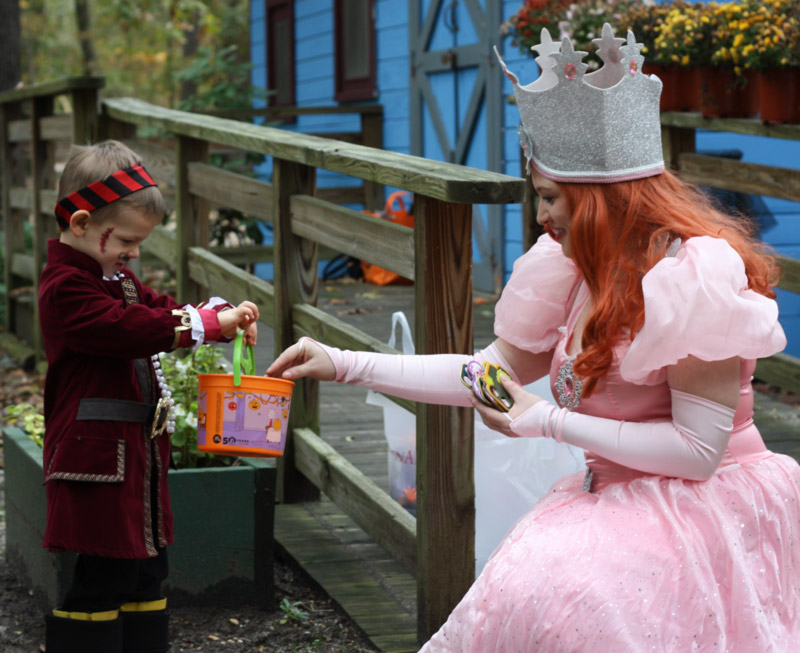 Image of a child dressed in a pirate costume holding a patterned Halloween Trick-Or-Treating basket with a light green handle toward a HalloWheaton performer outside in front of the blue WheatonArts Paper Studio. The performer has long red hair and is wearing a light pink gown with puffy sleeves, a large silver crown, and long light pink gloves. The performer is placing a HalloWheaton Trick-Or-Treating prize into the child's bucket.