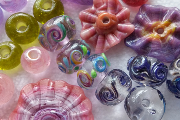 Image of an assortment of beads from Chris Coffman. There are two small light purple beads, three light small green beads, two small light pink beads, three clear beads with multicolor swirls in the middle, four clear beads with blue swirls around the middle, and three flower-shaped pink and purple beads with orange centers.
