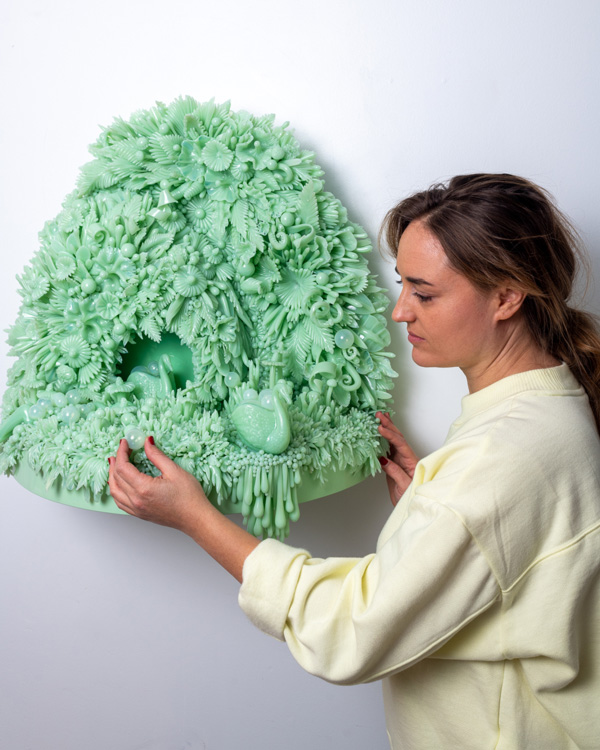 Photo of Amber Cowan standing to the right of and facing a mint green piece of her glass art. The piece is mounted on a white wall and is comprised of small pieces of mint green glass in the shape of various things from nature such as flowers, leaves, and mushrooms. The pieces make up the background and ground of the piece. Two larger mint green glass frogs are placed on the ground of the piece. One frog is inside a mint green cave in the center. Amber has her brown hair tied back with some loose strands in the front and is wearing a cream long-sleeved sweater rolled up slightly on the arm. Amber is reaching to touch one of the glass pieces on the ground portion of the work.