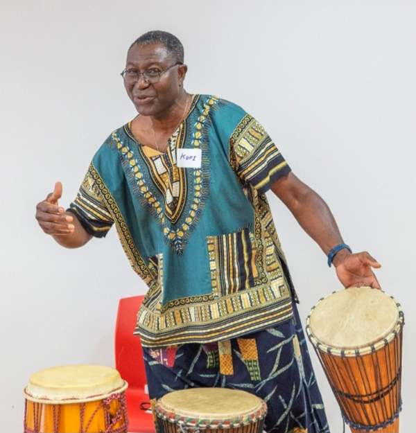 Image of Maxwell Kofi Donkor standing in front of a red chair and white background gesturing towards a drum that he is holding in his right hand. There are two other drums in front of him on the left side and middle of the image.