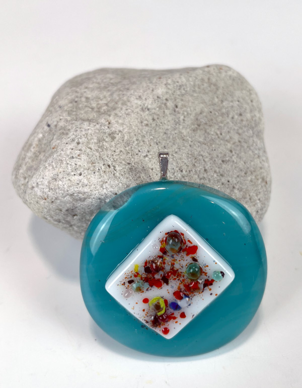 Image of a round blue fused glass pendant resting on the side of a light gray rock to hold it up in front of a white background. The pendant has a white rectangular piece of glass in the center with red, blue, and yellow speckles and small blue and green round attachments throughout.
