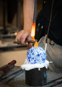 Image of a blue and white frit ornament being created in the WheatonArts Glass Studio. The image is a close up of the ornament resting a short, wide, and black cylindrical stand with white cloth at the top. There are various glass sculpting tools resting around the stand on top of the bench that the stand is on. A member of the WheatonArts Glass Studio team is adding the hook holder at the top of the ornament by pouring and shaping orange hot glass with a metal rod and pliers. One hand is pulling the hot glass away from the ornament and the other is squeezing the pliers to shape the hot glass into a hook.