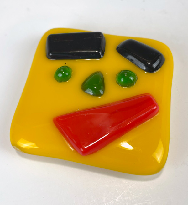 Image of a yellow square fused glass magnet in front of a white background. The magnet has two black rectangular glass eyebrows, two small round green eyes, and a small green triangular nose. The piece also has a red slanted rectangular mouth that is small at the top and gets larger towards the bottom in the bottom right corner.