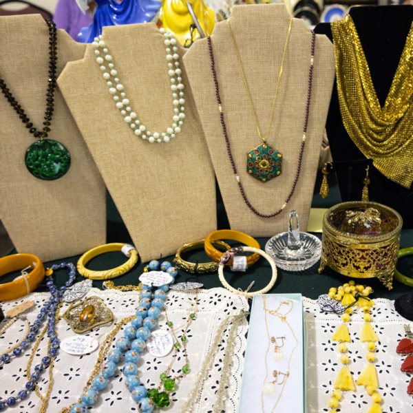 Image of antique jewelry at the WheatonArts Mid-Winter Antique Show. There are five necklaces resting on necklace stands. The pendant on the left is round with various shades of green and a black beaded chain. The necklace next to it is a two layer white beaded necklace with alternating large and small white beads. Next to that necklace is a green hexagon-shaped pendant with red and gold dots in a floral like pattern on a gold chain. The necklace around it has small purple beads and five visible longer silver beads to separate the purple beads into even sections. The necklace on the right is a gold triangular wrap with a gold tassel that has a gold bead at the top. Below the necklace stands are five bracelets. Three are yellow, one is gold, and the other is white. A clear ring holder and a gold antique jewelry box sit next to them on the right side of the image. Below those are more necklaces resting on white square doilies. The necklaces are made up of various sizes of purple, blue, yellow, red, green and gold beads and chains. One also has a gold diamond shaped pendant with a gold jewel in the middle.