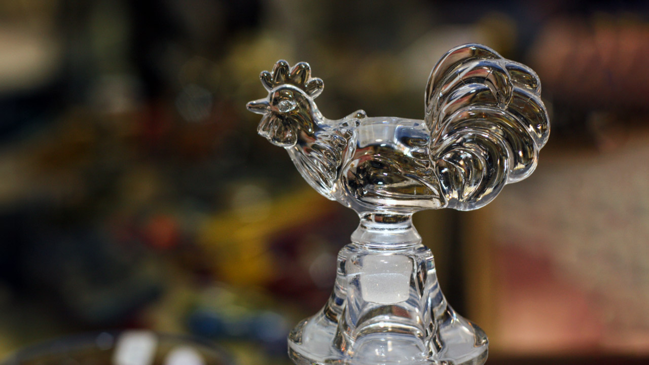 Close up image of an antique clear glass rooster attached to the top of a clear glass piece. The background is blurred.