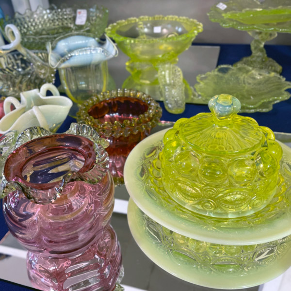 Image of antique glass vases and dishes at the WheatonArts Mid-Winter Antique Show. There are nine glass pieces shown. The pieces are various shades of purple, yellow, white, and clear glass shades.