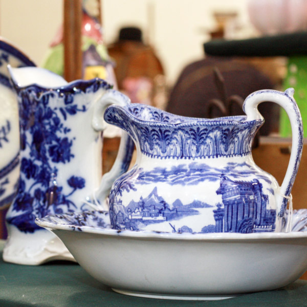 Image of two antique creamers and plates at the WheatonArts Mid-Winter Antique Show. The pieces are white with blue floral designs. One creamer also has the blue landscape of a city along with the blue floral details.