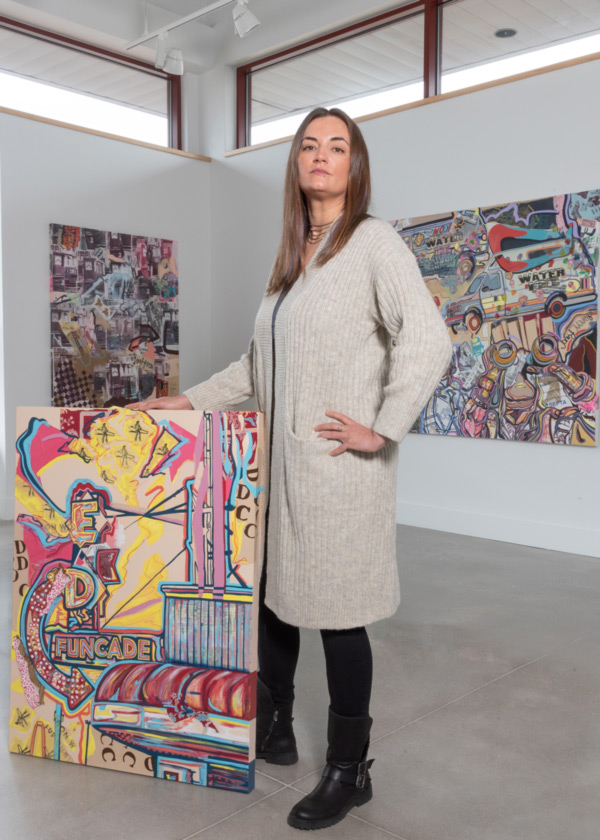 Image of Danielle Cartier standing in front of two white walls with a gray floor. Each wall has a piece of Danielle's artwork on it. Danielle has straight light brown hair and is wearing a long white long-sleeve cardigan over a black shirt and leggings. Danielle is also wearing black boots and has a hand holding up a piece of artwork. The artwork depicts the "Ed's Funcade" sign, building, and surroundings in a variety of yellow, red, blue, and purple neon colors.