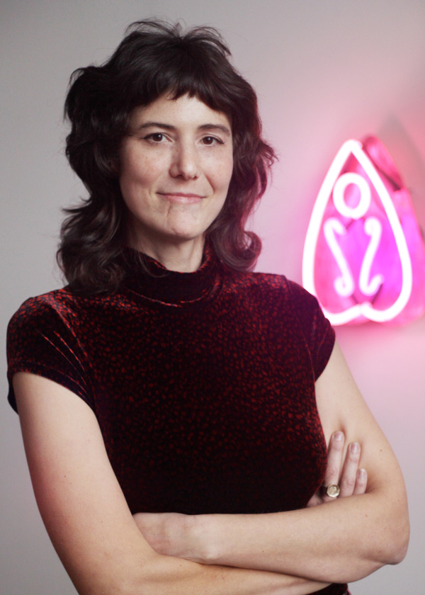 Headshot image of Alissa Eberle. Alissa had shoulder-length dark hair and is wearing a dark red shirt with short sleeves and a slight turtle neck. Alissa is standing in front of a white wall with a pink neon piece on the right side of it. The piece is shaped like a tall upside-down heart with a pink neon circle in the middle under the point. Below the circle are two curvy neon pink strands next to one other.